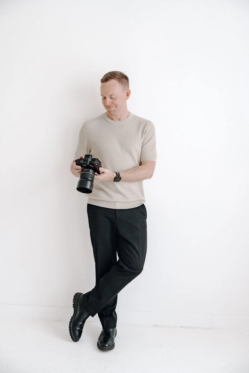 Portrait of James from Just James Weddings standing against a clean white studio wall, looking down at his camera while holding it in both hands, for his Somerset wedding photography website and personal about page introducing the photographer.