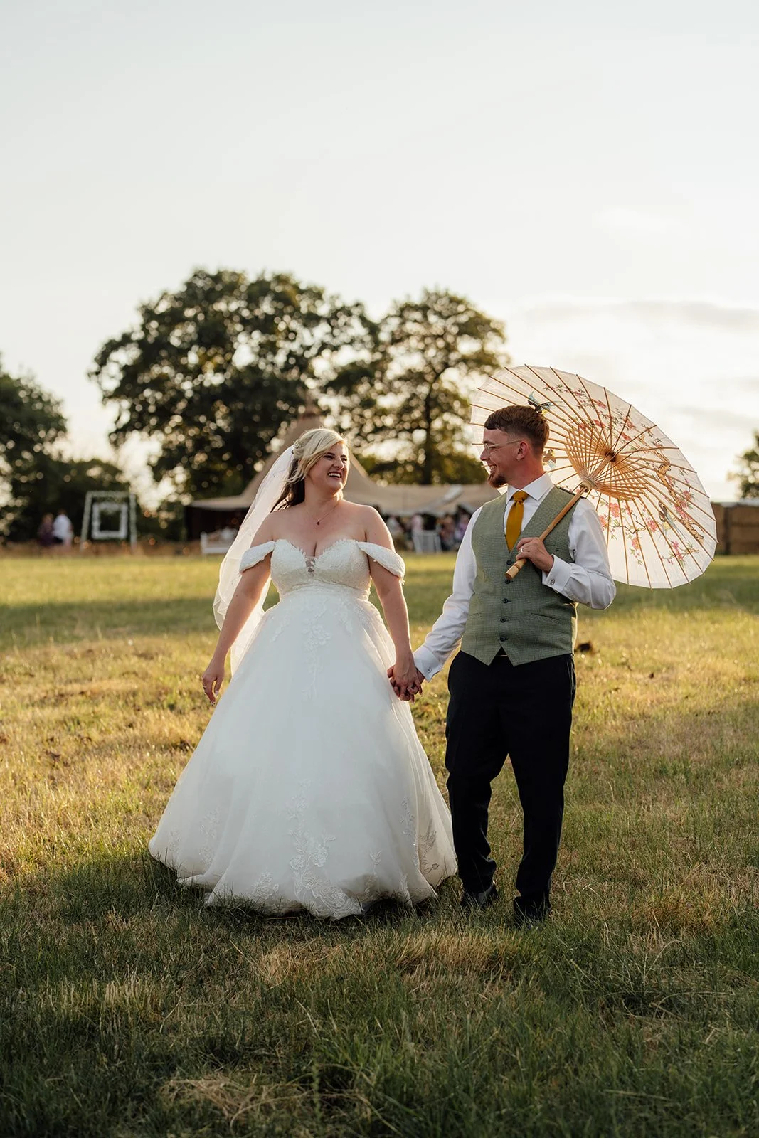 Lucy and Matthew walking hand in hand through the field at golden hour, with Matthew holding a parasol at Curated Canvas at Noah’s Pond in Somerset, photographed by Just James Weddings.
