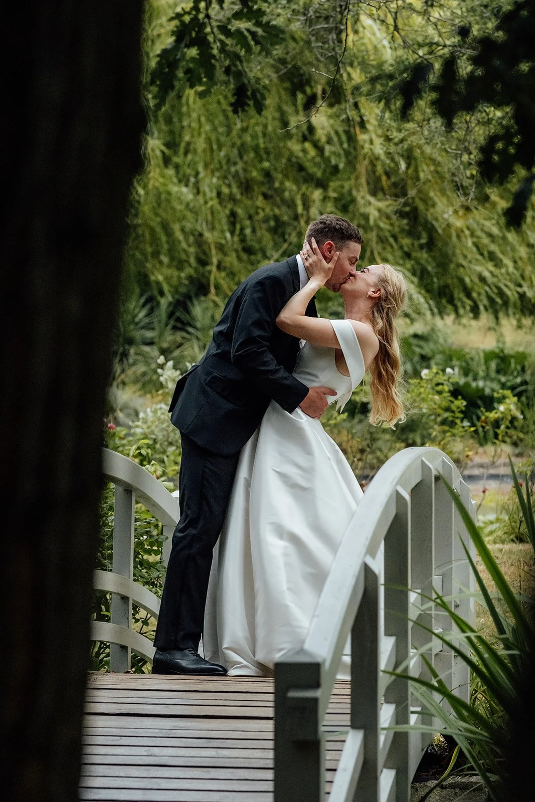Caitlin & Kevin sharing a kiss on a garden bridge at Clevedon Hall in Somerset, surrounded by greenery and water, photographed by Just James Weddings.
