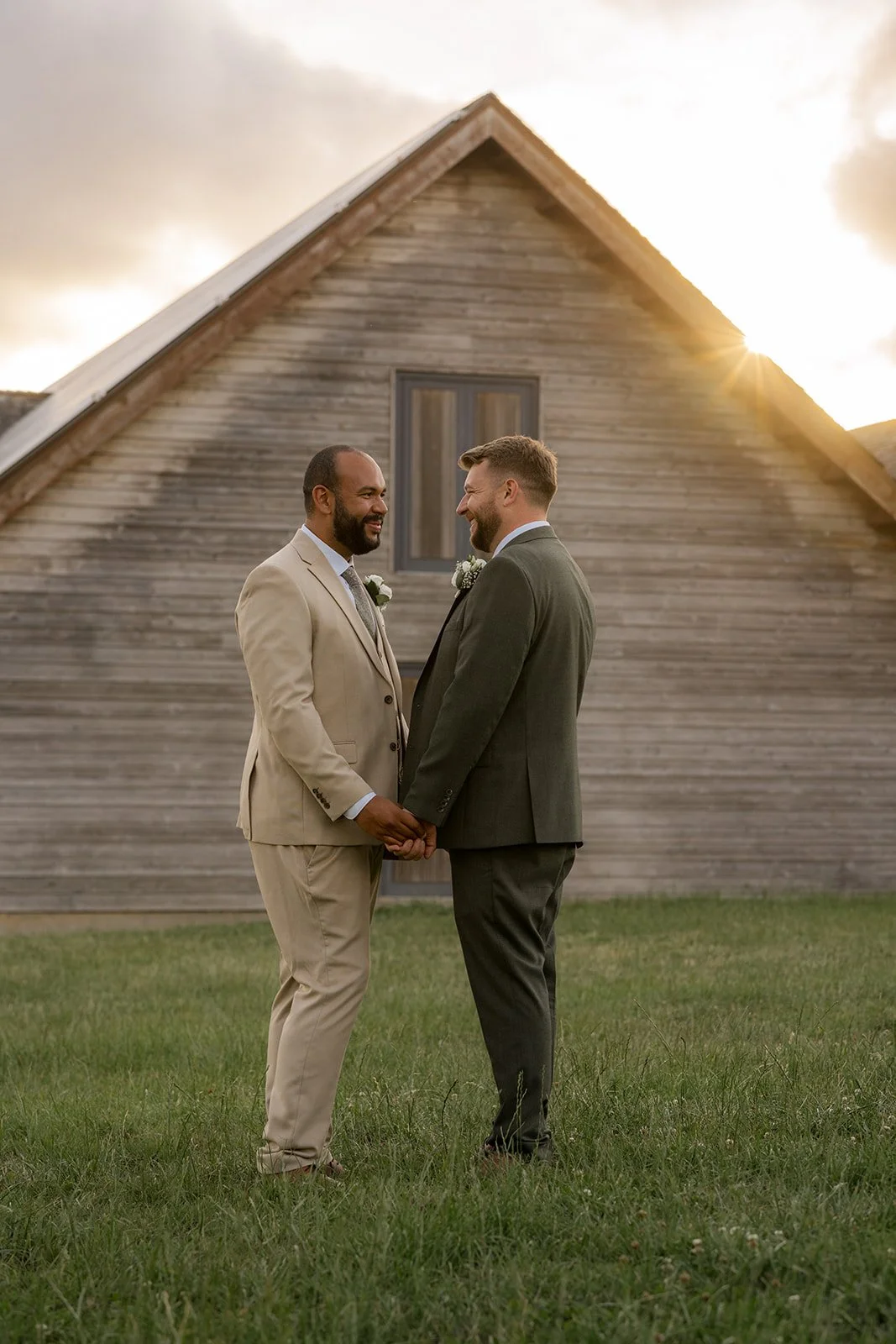 Aaron & Jack smile at one another while holding hands in the evening light outside Rock Village Hall in Worcestershire, photographed by Just James Weddings.