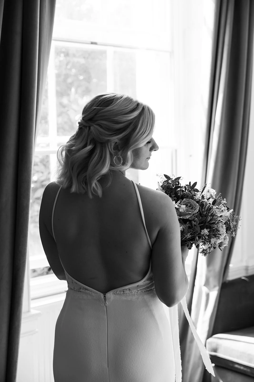 Black and white bridal portrait of Kristy holding her colourful wedding bouquet by a window at No. 4 Clifton Village in Bristol, UK, showing the back of her elegant wedding dress.