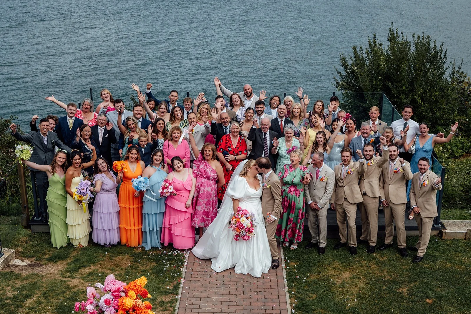 Holly & Alex kiss in front of their cheering wedding guests during a large group photo overlooking the sea at Sandy Cove Hotel in North Devon, photographed by Just James Weddings.