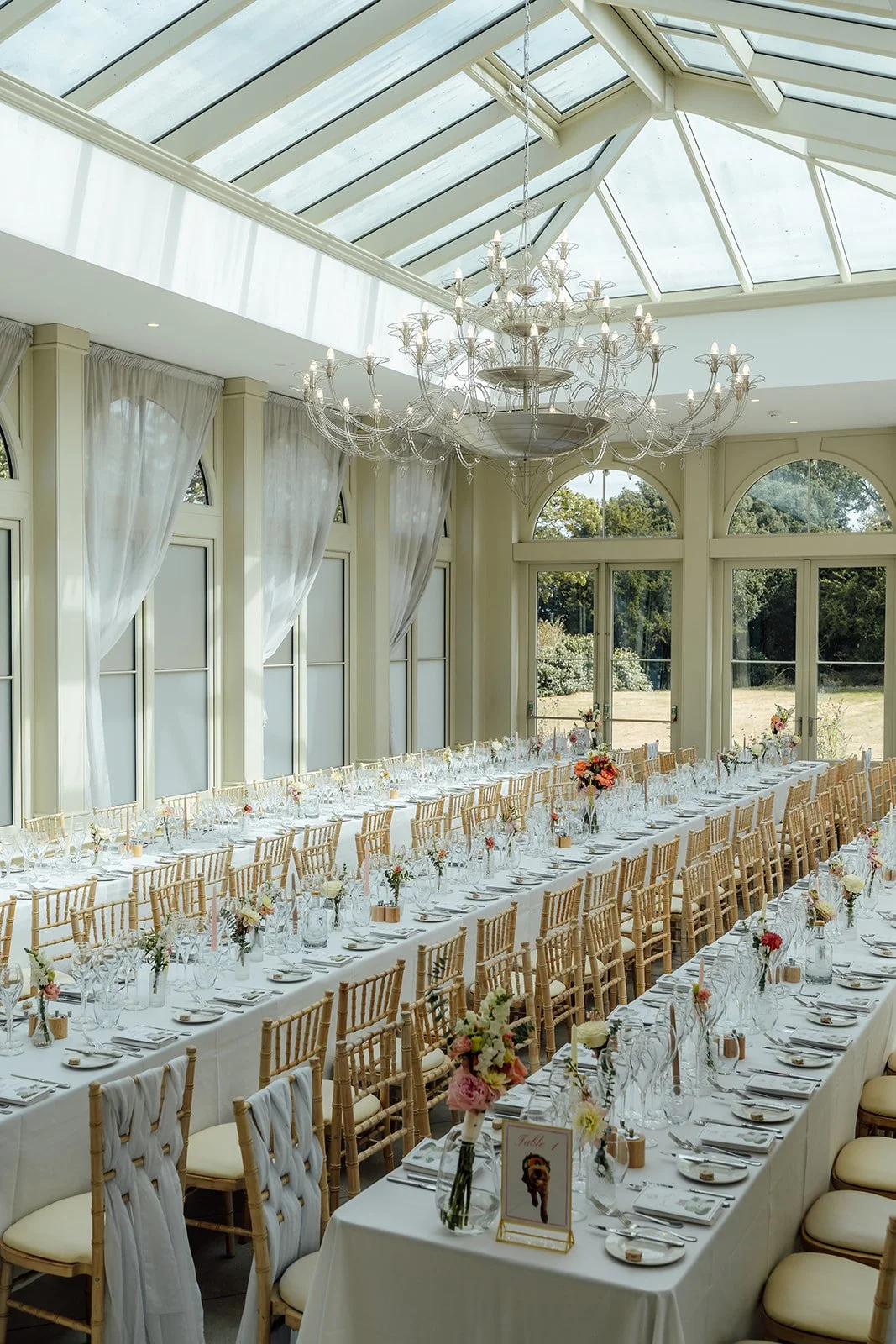 Bright wedding reception room at Clevedon Hall in Somerset, with long tables, gold chairs, floral details and a glass ceiling, photographed by Just James Weddings.
