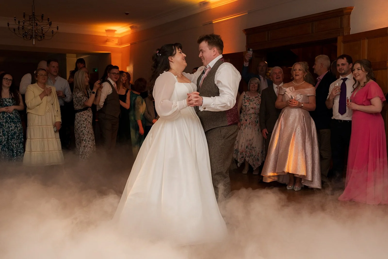 Emily & Josh share their first dance at Coombe Lodge Blagdon, Somerset, UK, surrounded by low-lying fog and wedding guests watching around the dance floor. Evening reception photo with soft lighting, warm tones and the couple smiling at each other.