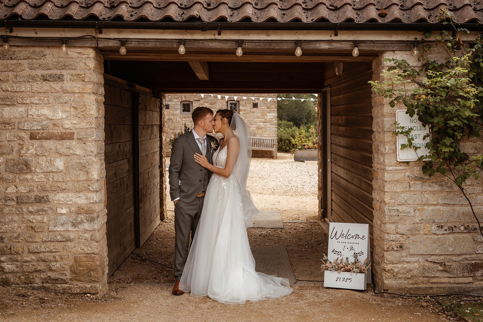 Madeleine & Izaac share a kiss beneath a rustic stone archway during their wedding at Folly Farm Centre in Somerset, UK, surrounded by warm textures, hanging lights, and countryside details that frame their intimate portrait moment.