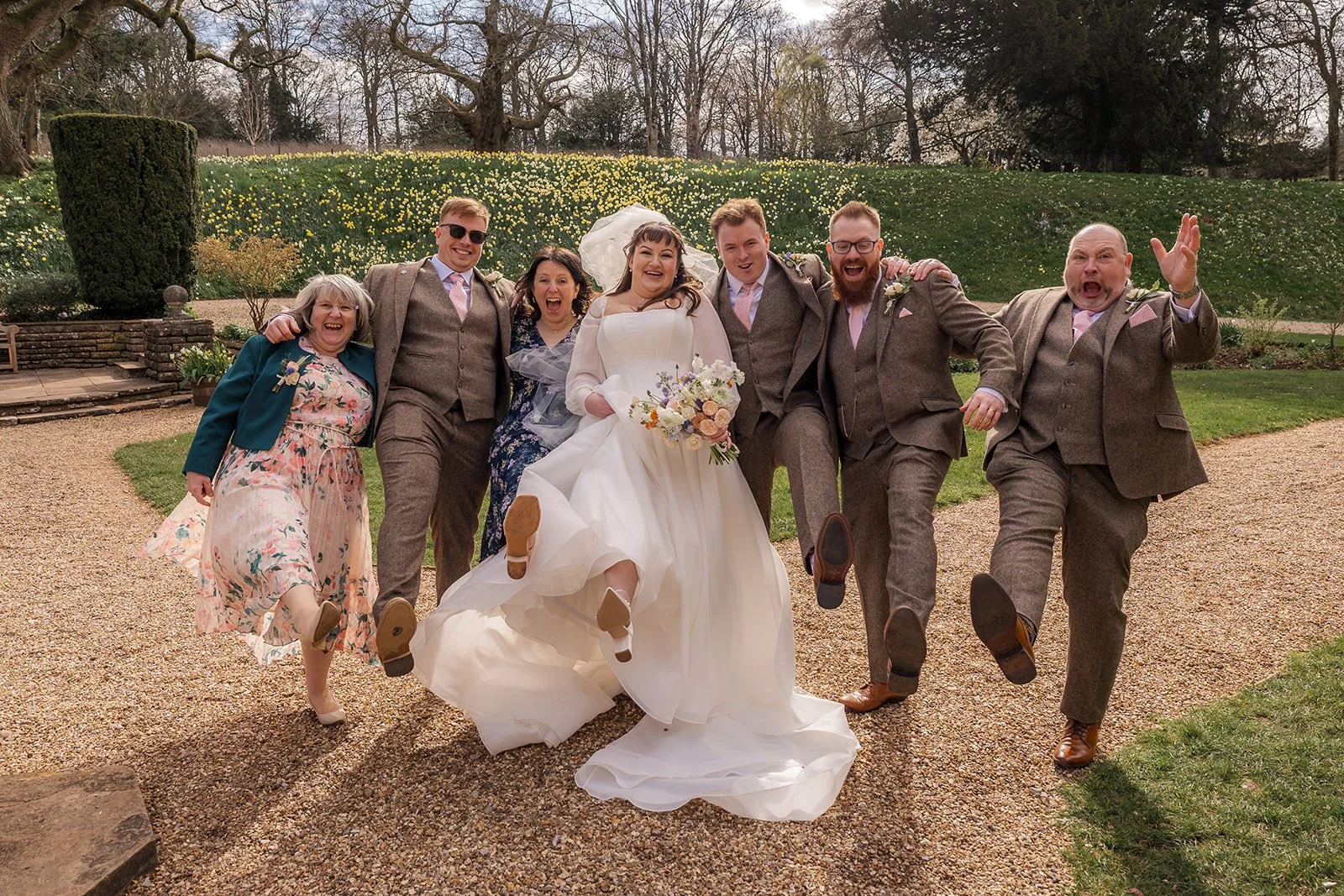 Emily and Josh pose with friends and family in a fun wedding group photo at Coombe Lodge in Somerset, UK, with everyone kicking their legs up in celebration.