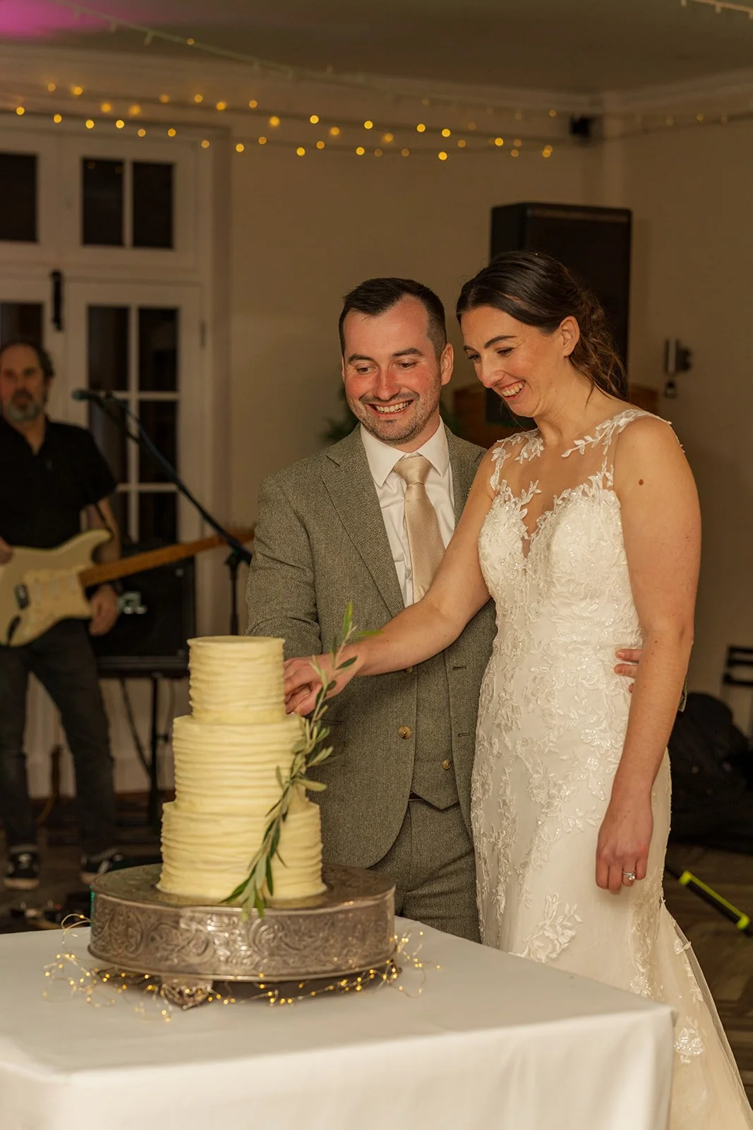 Anna and Chris cut their wedding cake at Elmhay Park in Somerset, UK, smiling together beside a tall ivory cake as fairy lights glow overhead and the band waits behind them. A joyful evening reception moment from Anna & Chris’s wedding day.