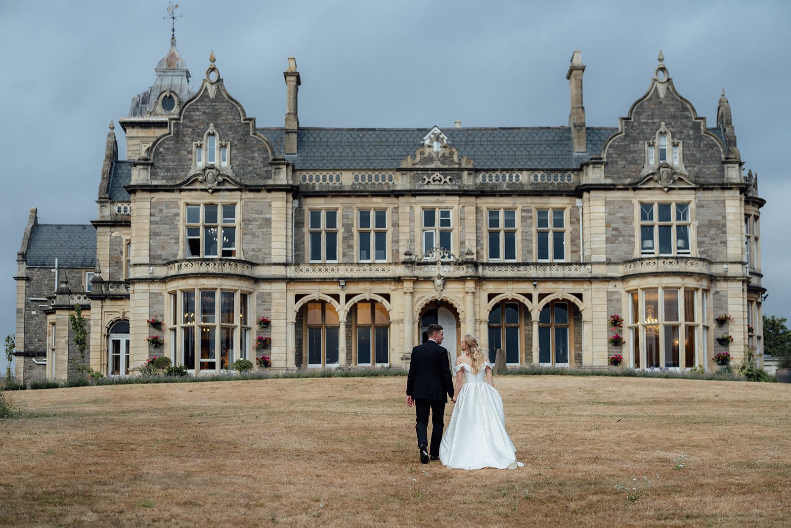 Caitlin and Kevin walking hand in hand across the lawn towards Clevedon Hall in Somerset, with the grand historic wedding venue filling the background.