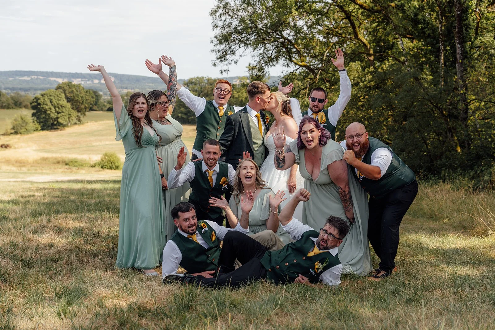 Lucy and Matthew kissing in the centre of their cheering wedding party during a fun countryside group photo at Curated Canvas at Noah’s Pond in Somerset, photographed by Just James Weddings.