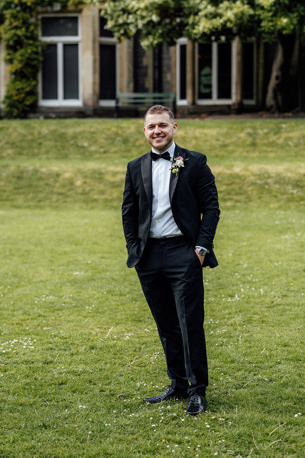 Max smiles for a solo groom portrait on the lawn at Insole Court in Cardiff, wearing a black tuxedo with boutonniere, hands in pockets and the venue’s historic stone house softly blurred behind him after Bryony & Max’s ceremony in the gardens.