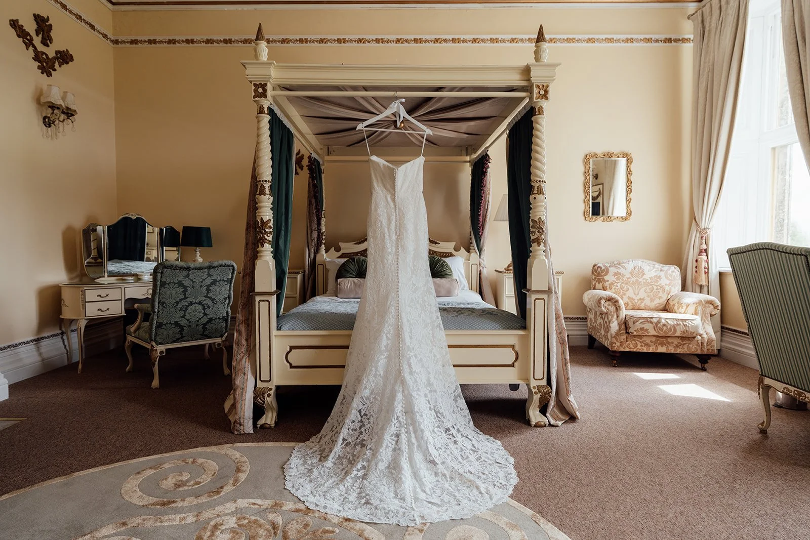 An elegant lace wedding dress hanging from a four poster bed in a grand bridal suite at Orchardleigh Estate in Somerset for Amy & Matt’s wedding morning hero shots by Just James Weddings.