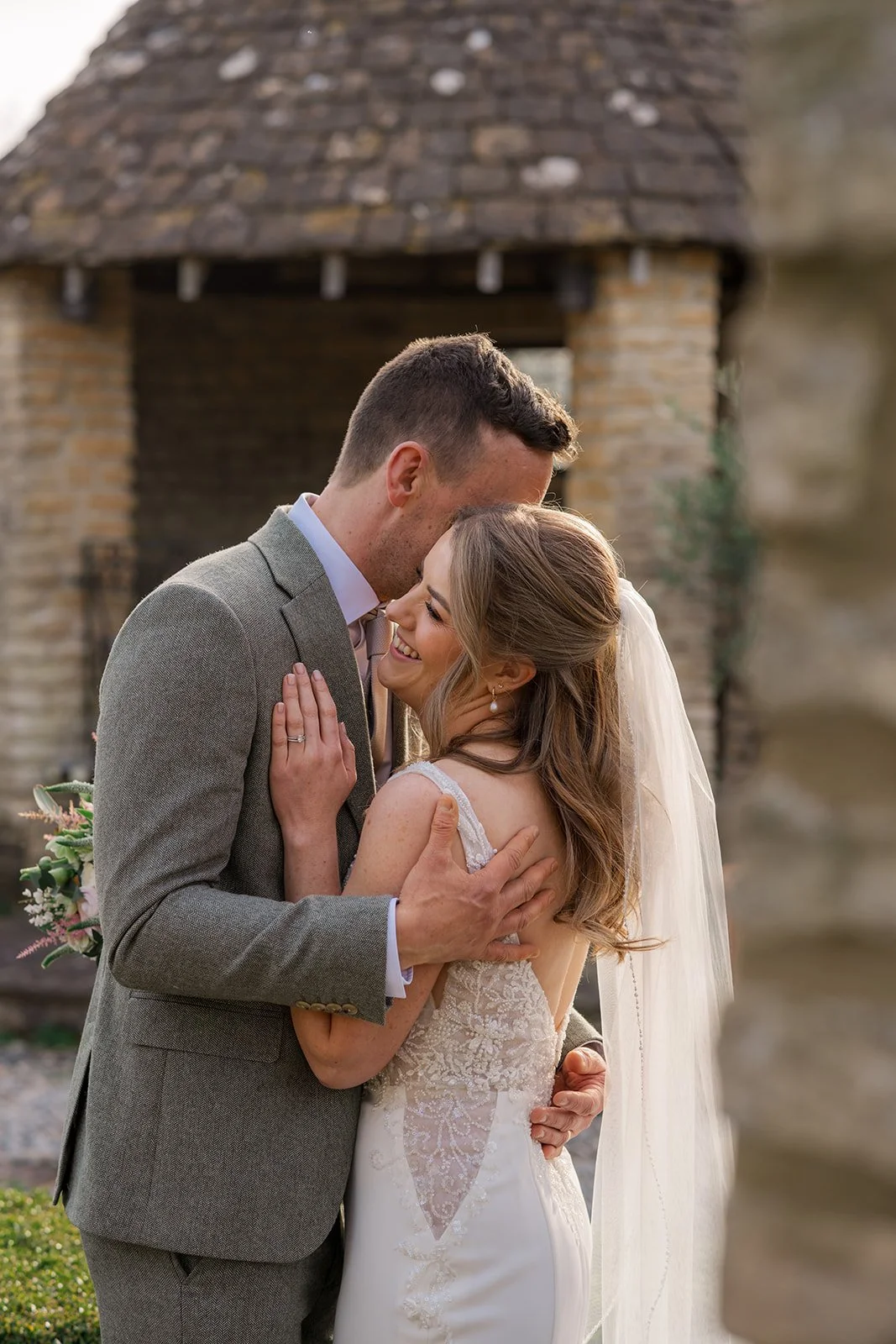 Anna and Tom embrace during wedding portraits at Winkworth Farm in the Cotswolds, UK. This close romantic image captures Anna smiling in her veil and lace gown as the couple cuddle in soft evening light during Cotswolds wedding photography.