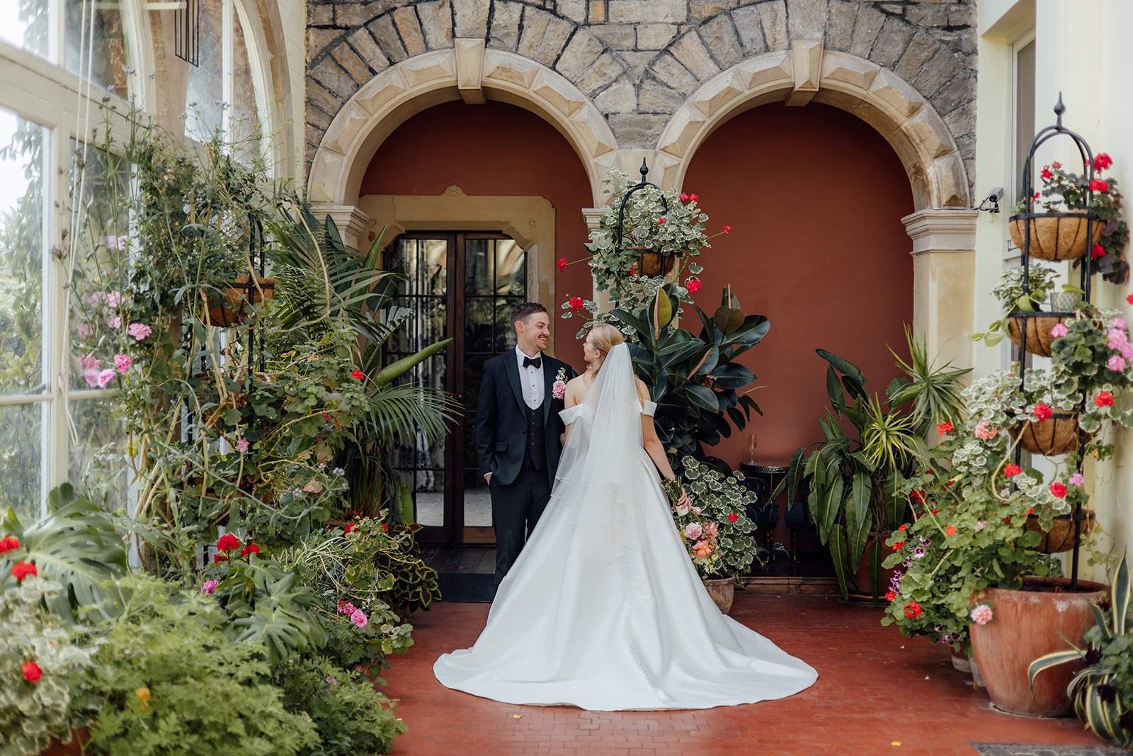 Caitlin and Kevin standing together beneath the stone arches and tropical plants at Clevedon Hall in Somerset, looking at each other with Caitlin’s wedding dress and veil flowing behind her.