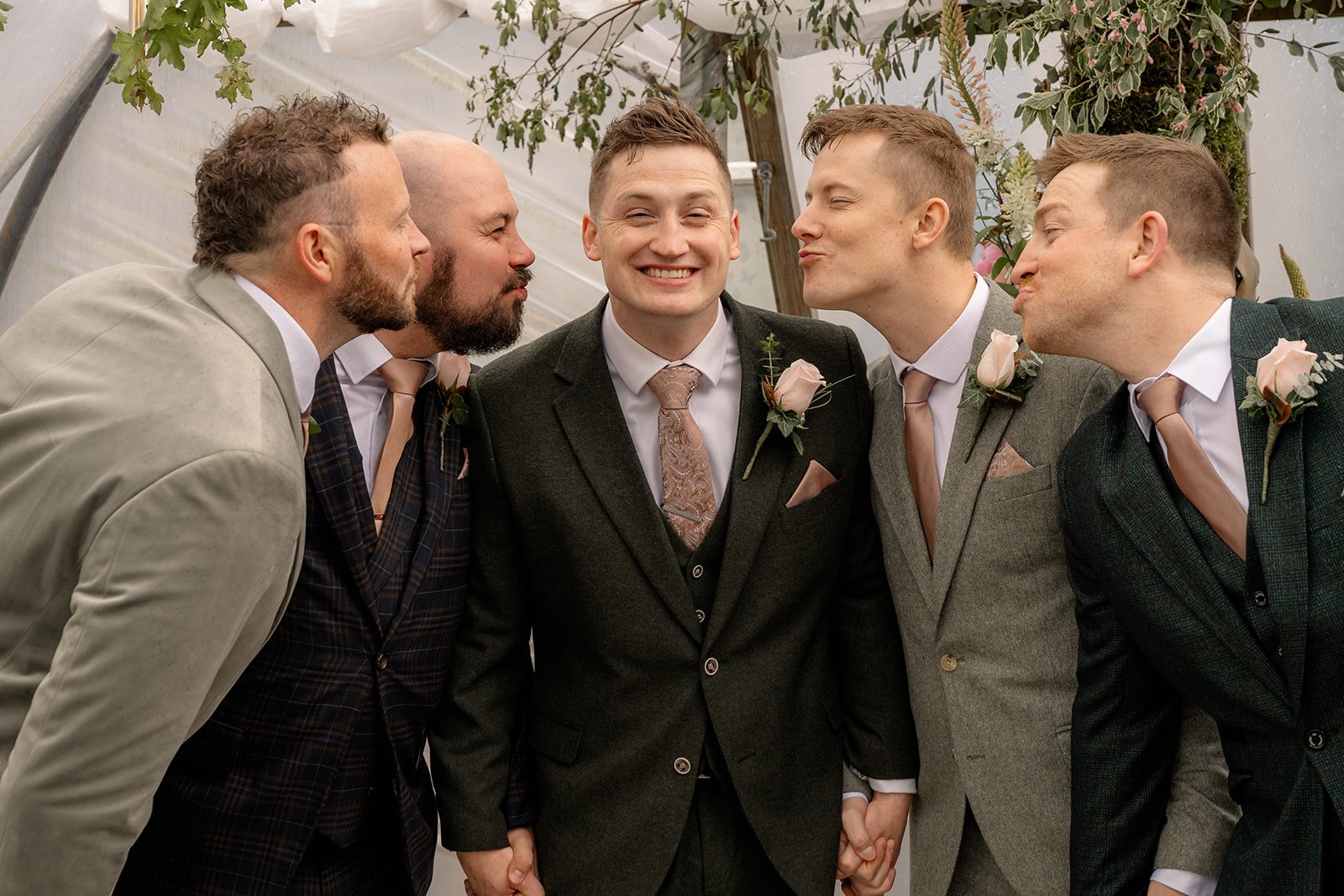 Jaye & Josh with the groom and groomsmen sharing a playful kiss pose during their Farrington's Farm wedding in Bristol, surrounded by soft greenery, pink buttonholes, matching ties and relaxed laughter inside the poly tunnel ceremony space.