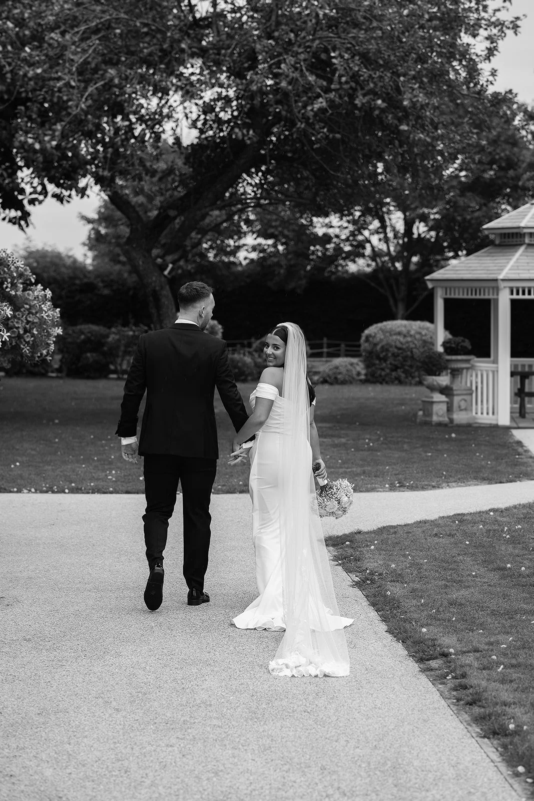 Nisha and Jack walk hand in hand through the gardens at Batch Country House in Somerset, UK, with Nisha looking back towards the camera as her long veil trails behind her beside the white garden gazebo, photographed by Just James Weddings.