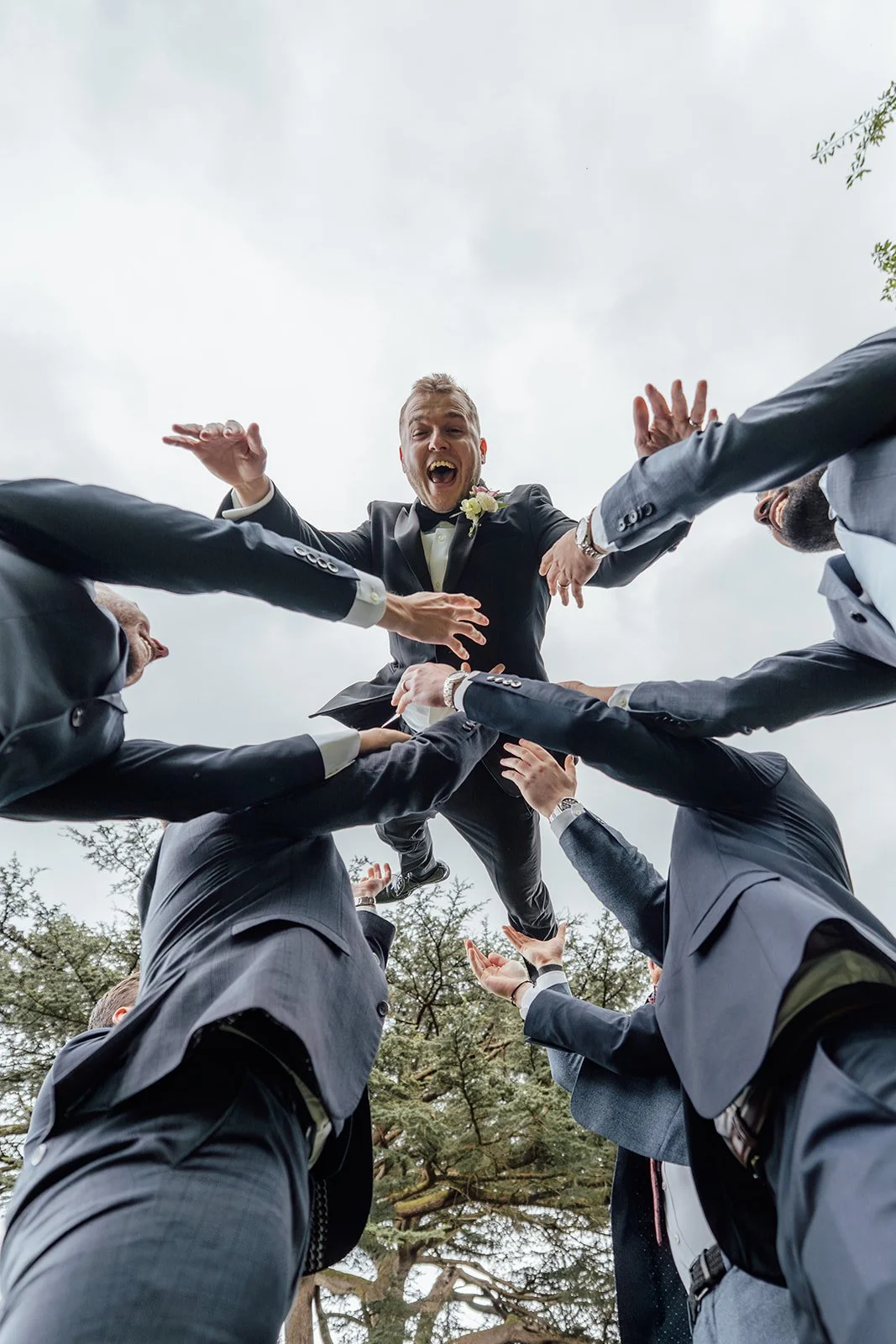 Max is lifted into the air by his groomsmen during Bryony & Max’s wedding at Insole Court in Cardiff, captured from a low angle with laughing faces, outstretched hands, smart suits and tall trees above the garden celebration outdoors together.