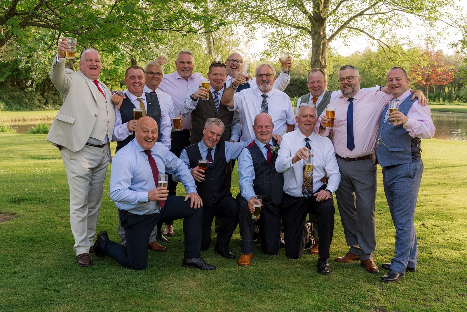 Debbie and Nigel’s wedding guests raise glasses by the lake at Yarlington Barn in Somerset, creating a lively group portrait with friends, sunshine and countryside gardens after their relaxed barn wedding ceremony.
