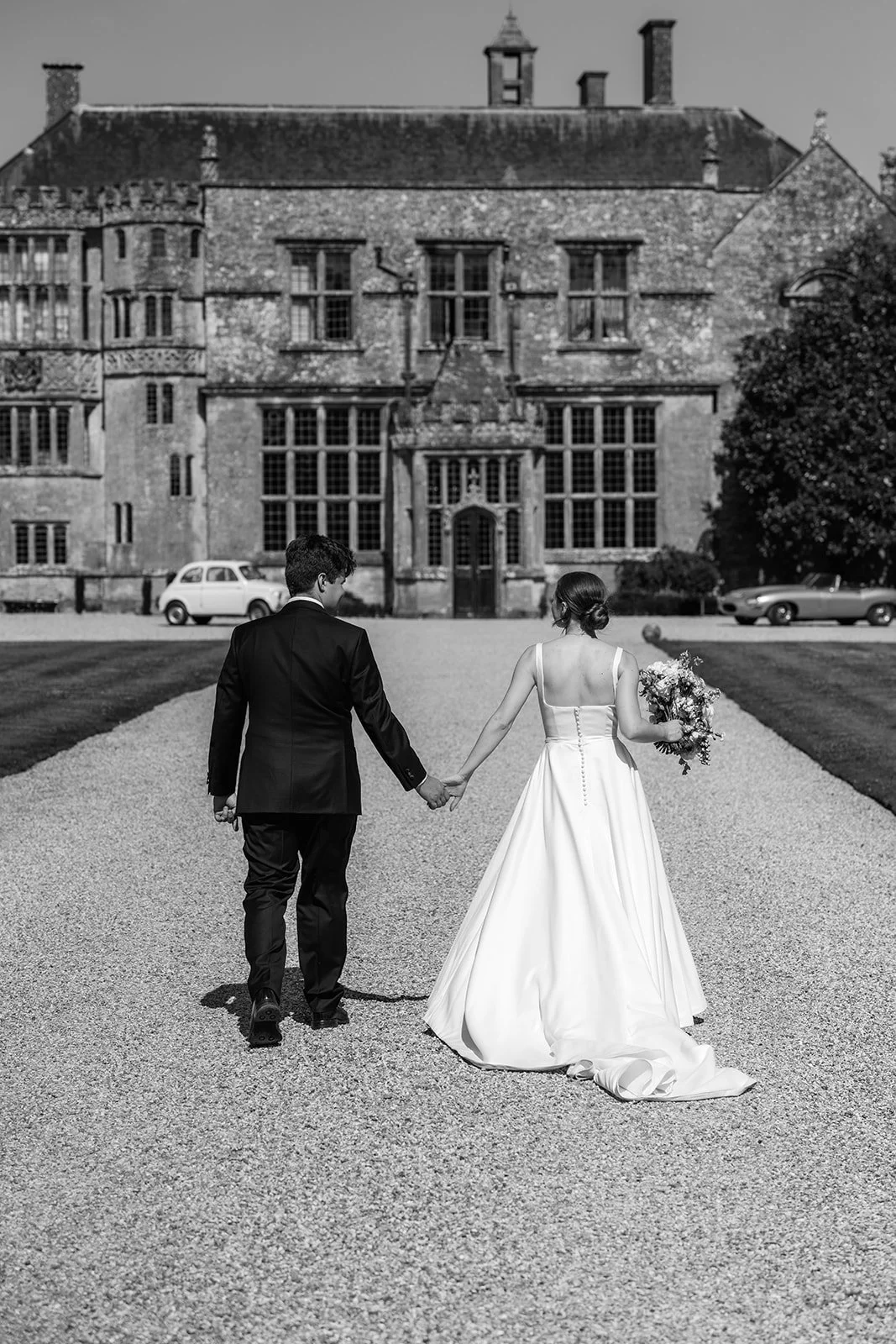 Black and white wedding photo of a couple walking hand in hand along the gravel driveway at Brympton House in Somerset, with the historic manor house ahead.