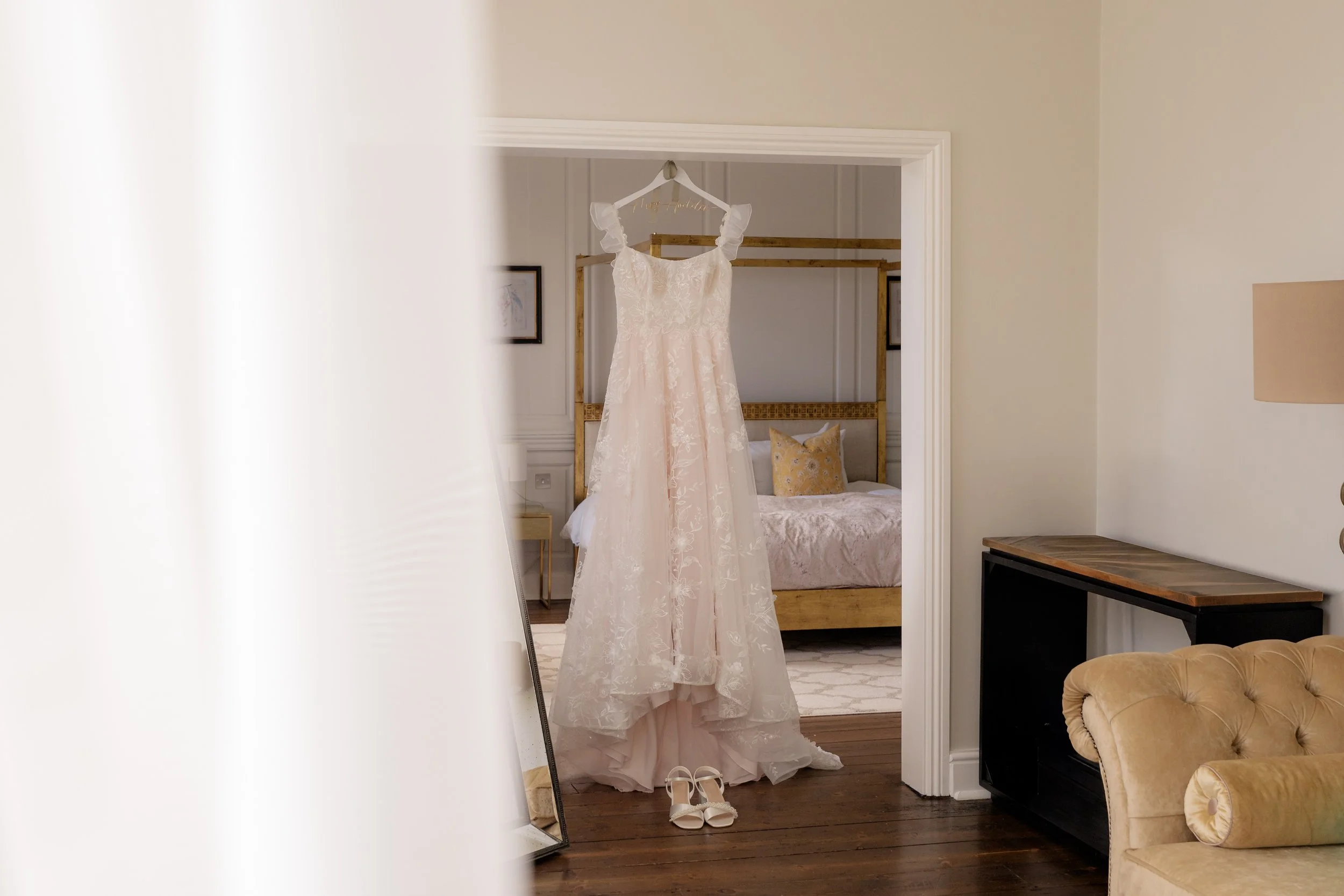 Jaye & Josh’s wedding at Syrencot in Wiltshire, featuring a delicate lace wedding dress hanging in a bright bedroom doorway with bridal shoes below, soft neutral interiors and elegant bridal preparation details before the ceremony began.