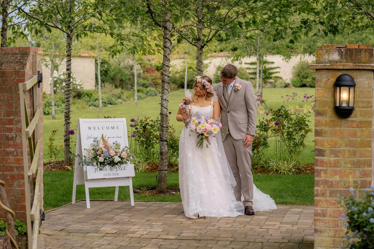 A bride and groom walking together outdoors during their wedding, surrounded by trees and flowers, with a welcome sign and floral arrangement nearby.