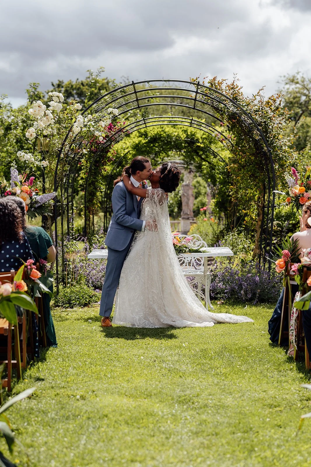 Melody & Jonny share their first kiss beneath a flower-covered garden arch during their outdoor wedding ceremony at Berwick Lodge in Bristol, photographed by Just James Weddings.