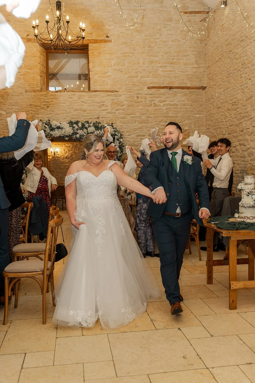 Siobhan & Nicholas walk into their wedding breakfast at Kingscote Barn in the Cotswolds, UK, holding hands and smiling as guests wave napkins around them, a lively rustic barn wedding reception entrance with stone walls, fairy lights and cake.