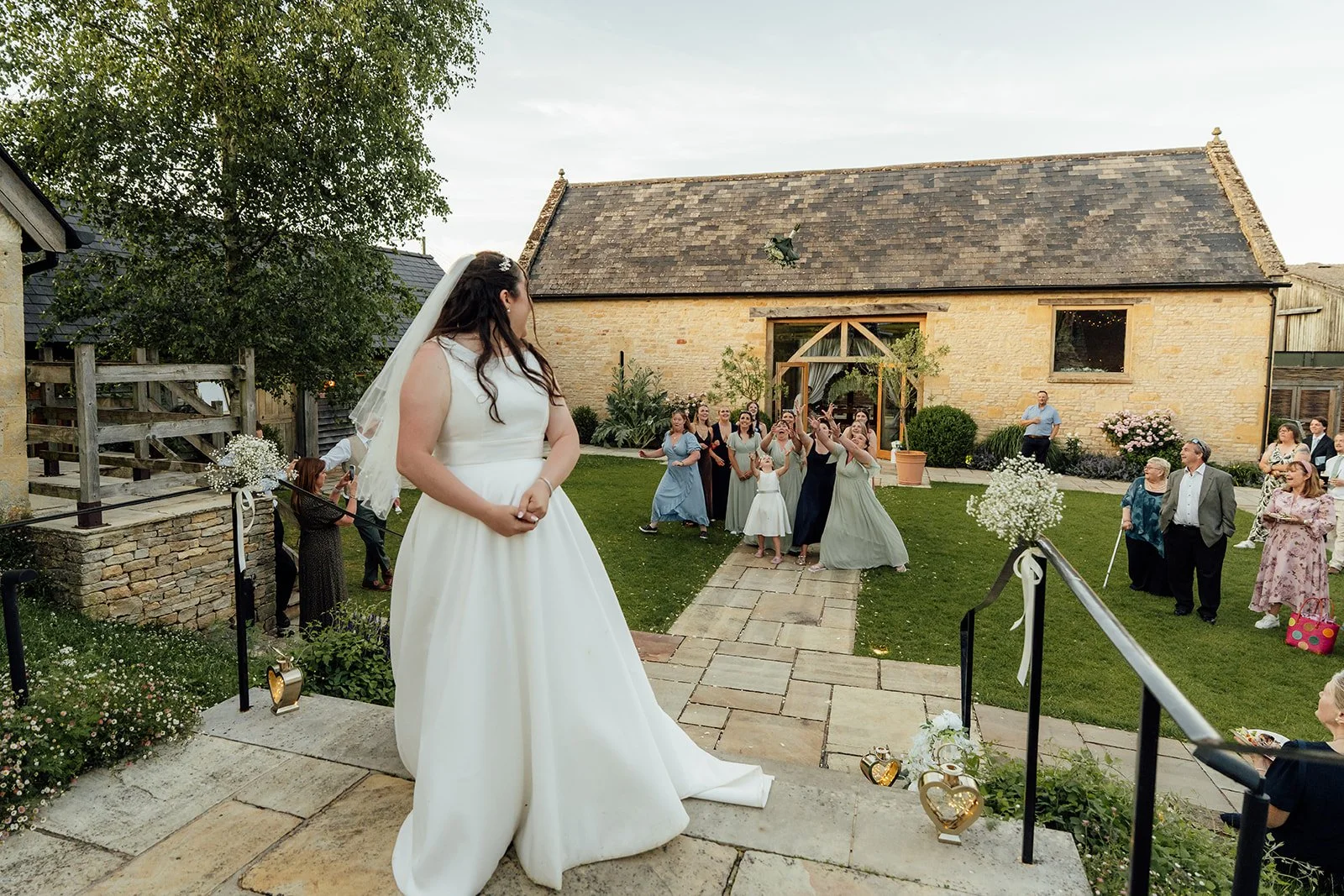 Chloe throws her wedding bouquet towards guests gathered in the courtyard at The Barn at Upcote in Gloucestershire during her wedding to Armani, photographed by Just James Weddings.