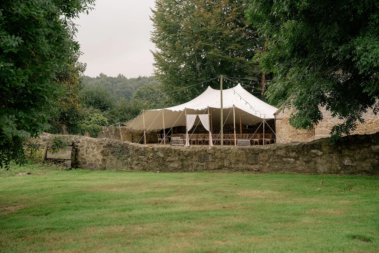 Outdoor ceremony marquee at The Cider Barn in Somerset for Abi & Josh’s wedding, with canvas canopy, stone wall, lawn and woodland around the setting, photographed by Just James Weddings for relaxed countryside wedding venue.
