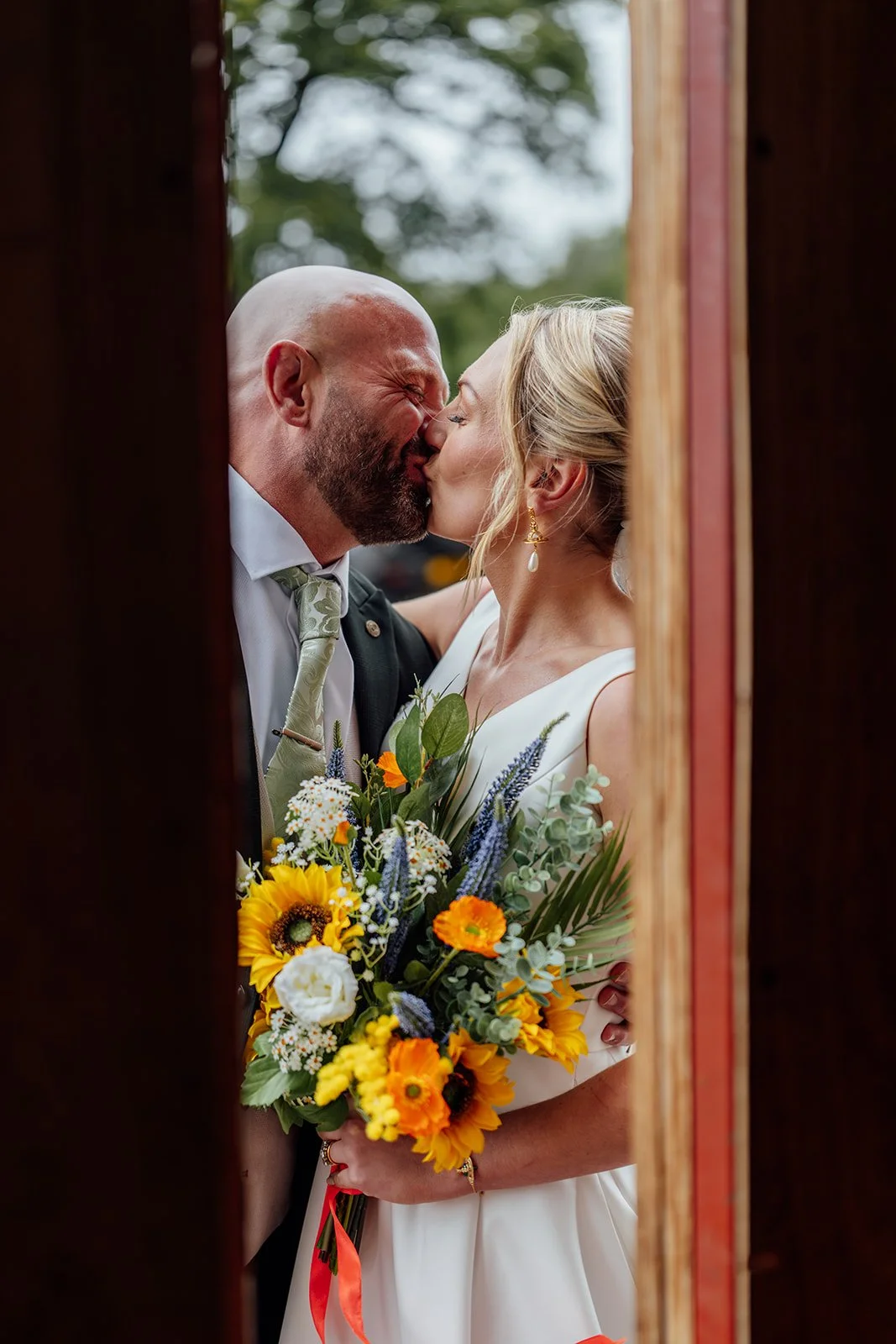 Close-up portrait of Hannah & Ben sharing a kiss at Grays Barn in Devon, UK, framed through wooden barn doors with her bright sunflower bouquet in the foreground, captured romantically and naturally by Just James Weddings after their ceremony.