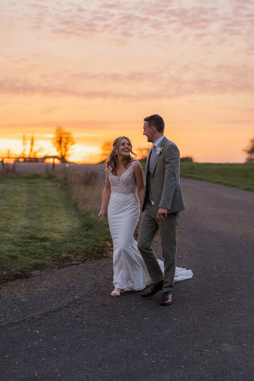 Anna and Tom walk together at sunset at Winkworth Farm in the Cotswolds, UK. This romantic wedding portrait captures the couple smiling on a country road beneath glowing skies, with soft evening light in timeless Cotswolds wedding photography.