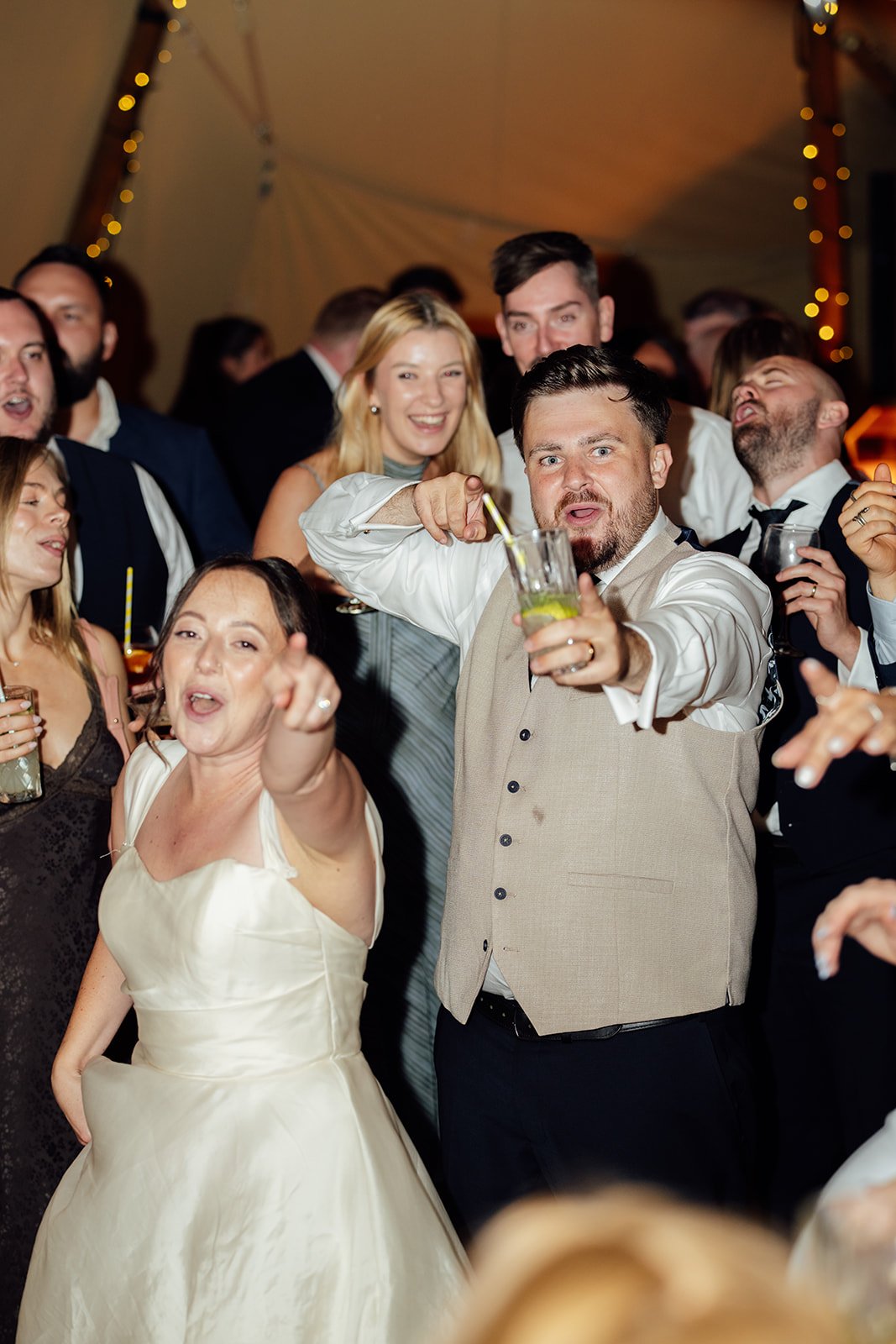 Harriet & Simon point towards the camera while dancing with friends inside the tipi at Dovecote Events in Oxfordshire, photographed by Just James Weddings.