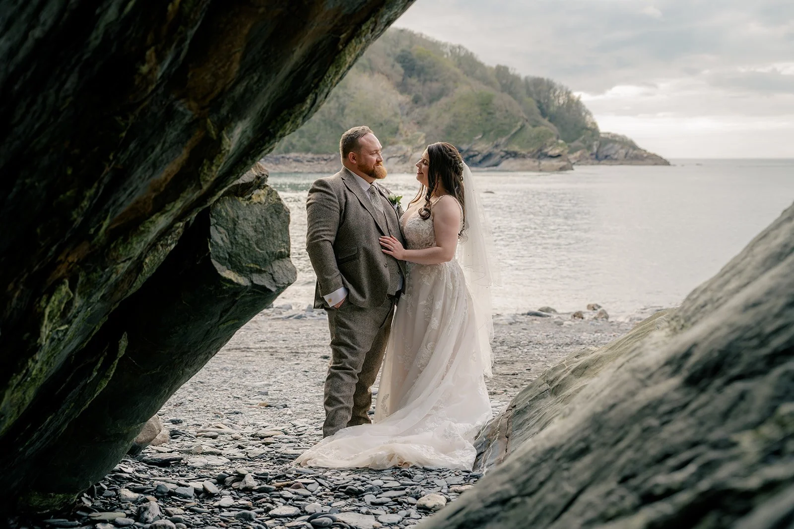 Sam & Terri standing together in a rocky beach cave at Sandy Cove Hotel in North Devon, with the bride in her veil and the sea behind them, photographed by Just James Weddings for romantic coastal wedding portraits and natural venue inspiration.