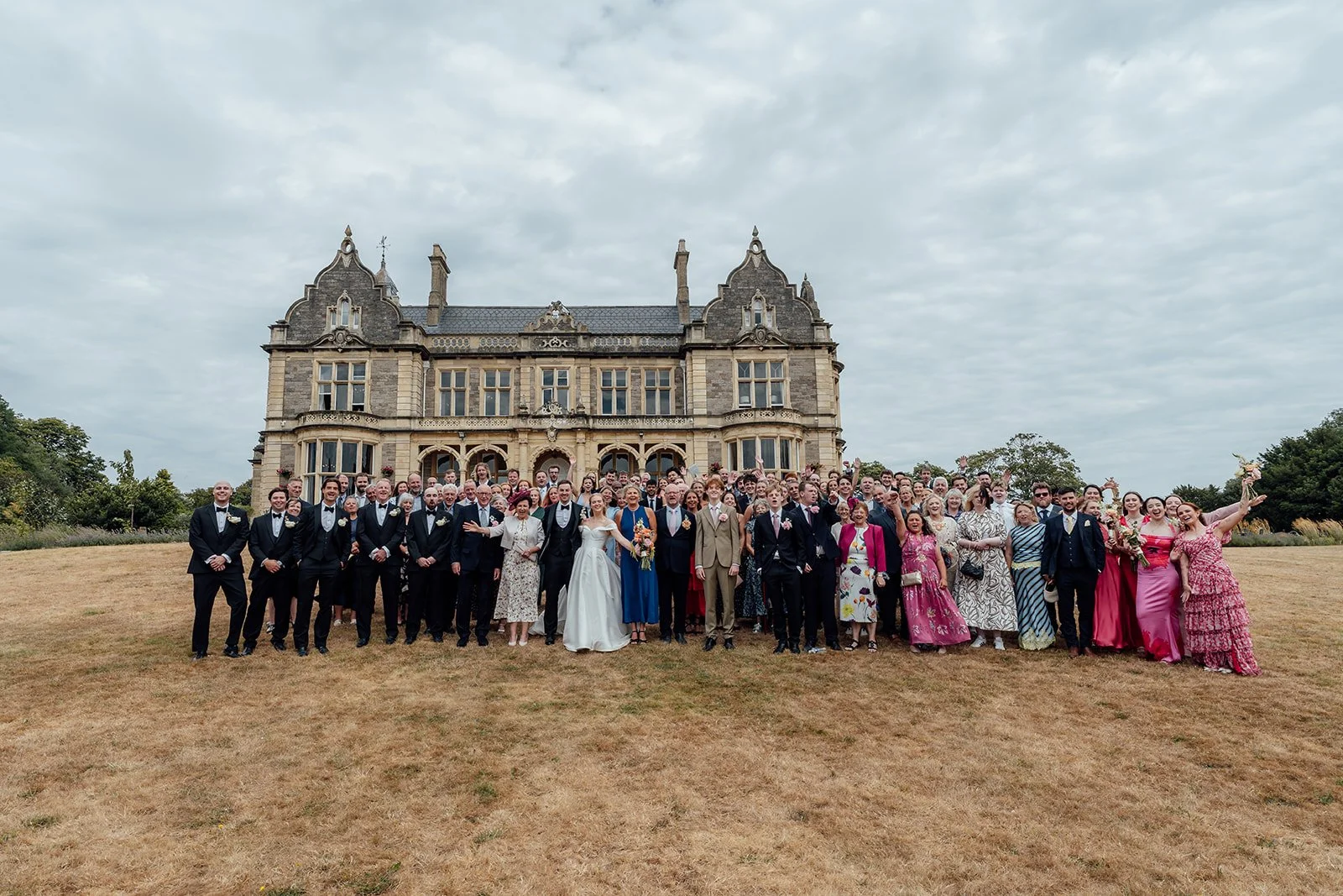 Large wedding group photo of Caitlin & Kevin with their guests on the lawn at Clevedon Hall in Somerset, with the historic manor house behind them, photographed by Just James Weddings.
