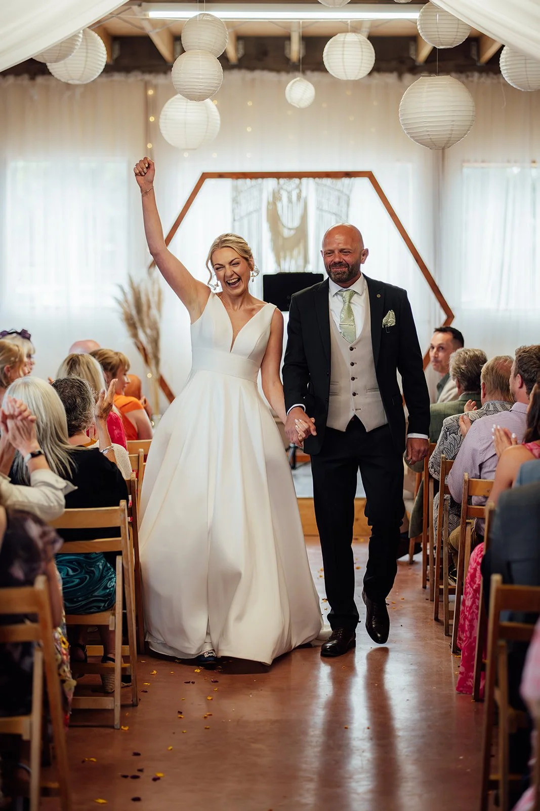Joyful aisle exit photo of Hannah & Ben at Grays Barn in Devon, UK, walking hand in hand after their ceremony as guests clap beneath paper lanterns and soft white draping, photographed with bright, celebratory emotion by Just James Weddings..