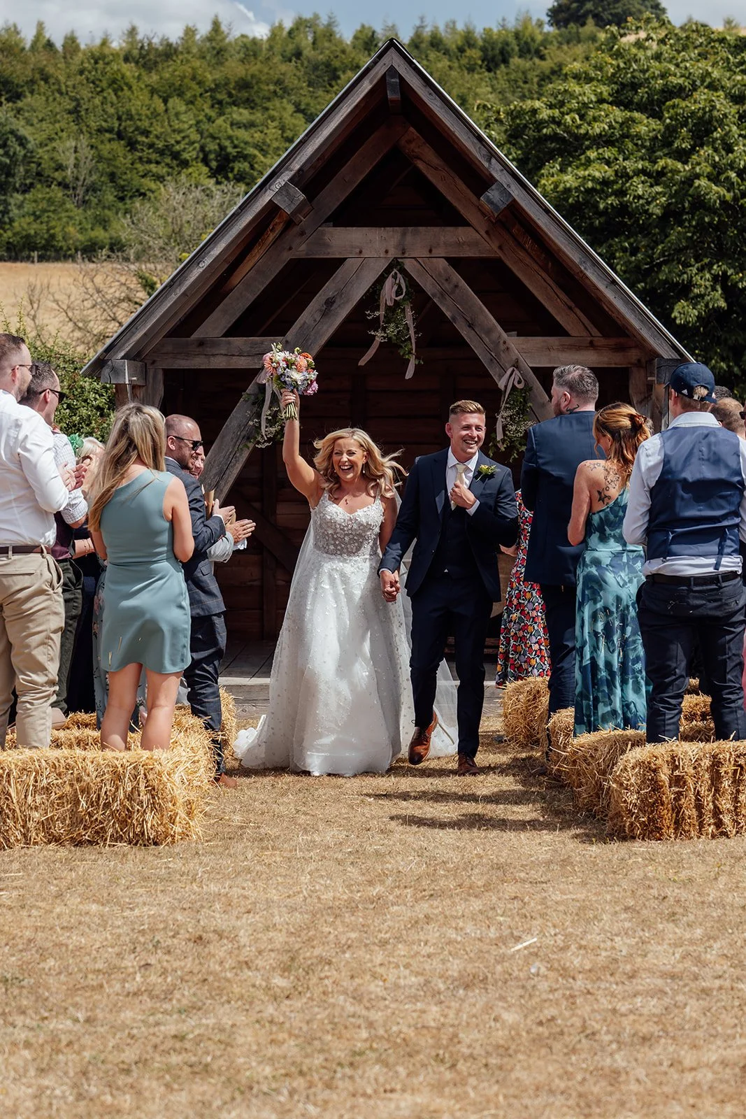 Elise and Tom walking hand in hand from their outdoor ceremony as guests cheer beside hay bale seating at Bowerchalke Barn in Wiltshire, photographed by Just James Weddings.