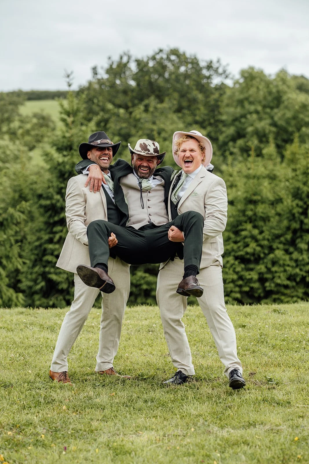 Playful groomsmen portrait from Hannah & Ben’s wedding at Grays Barn in Devon, UK, with two friends carrying Ben across the grassy countryside in cowboy hats, captured with fun, relaxed energy and laughter by Just James Weddings outdoors.