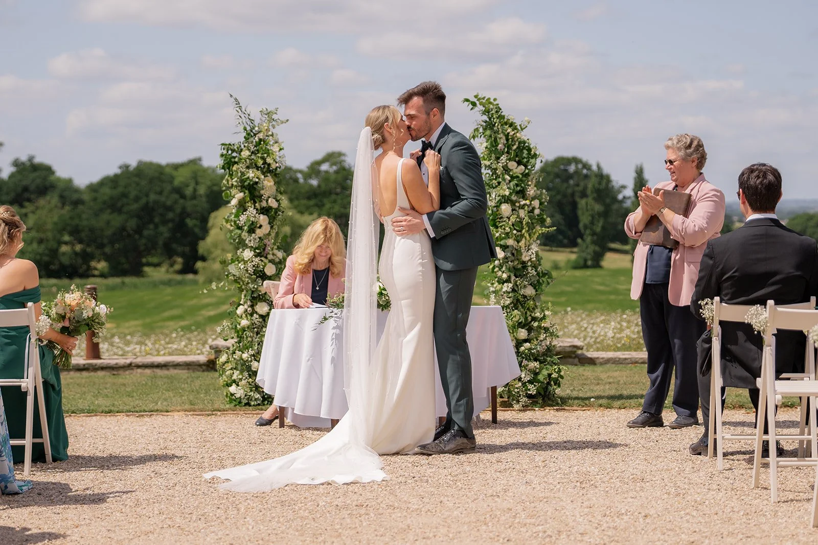 Chessie &amp; James at Rockbeare Manor sharing a kiss during their outdoor ceremony, standing between two floral pillars as guests watch and the registrar signs behind them. The scene is bright and sunny with views of the gardens and countryside.