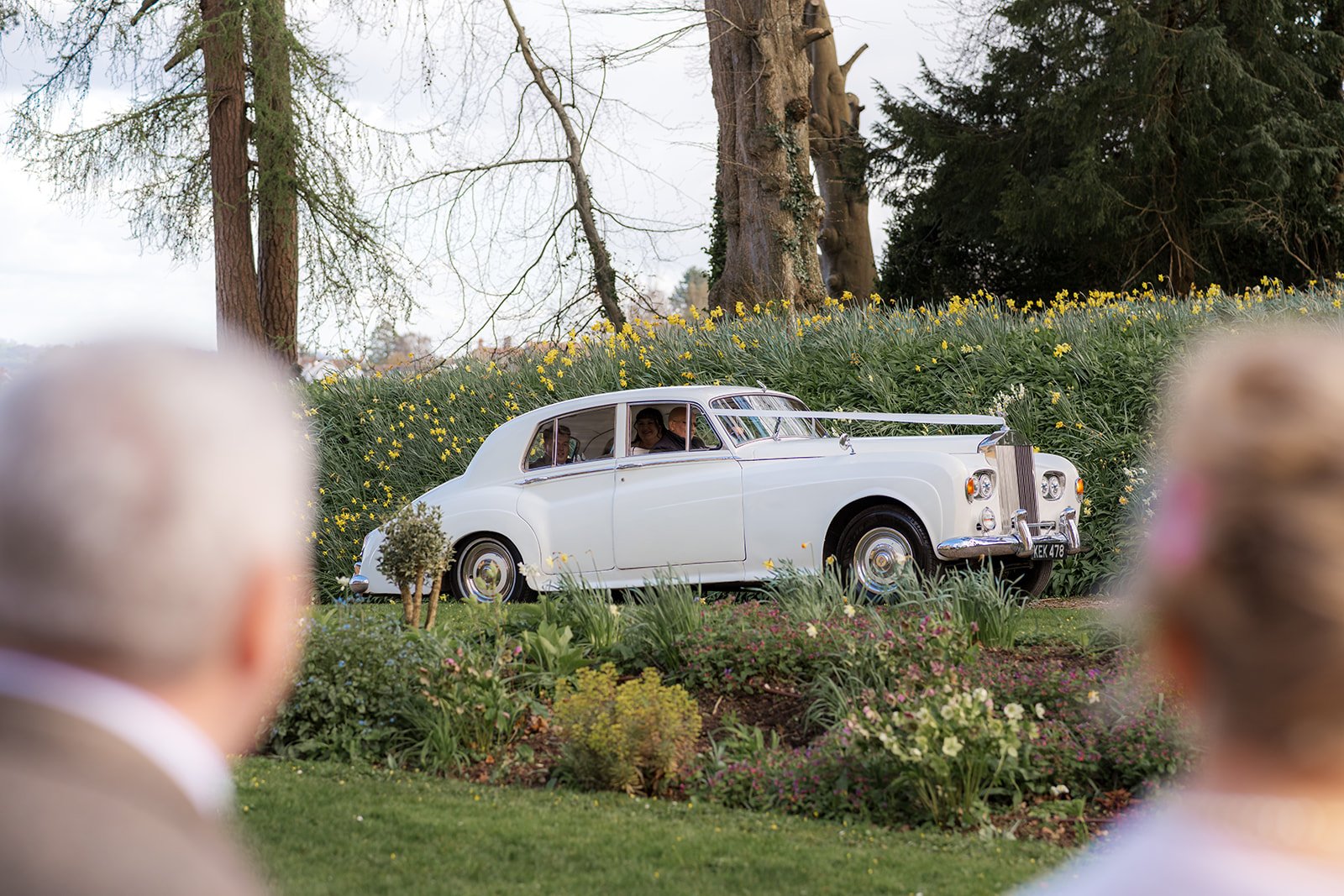 A classic white wedding car arrives through the gardens at Coombe Lodge in Somerset, UK, surrounded by spring flowers before Emily and Josh’s wedding ceremony.