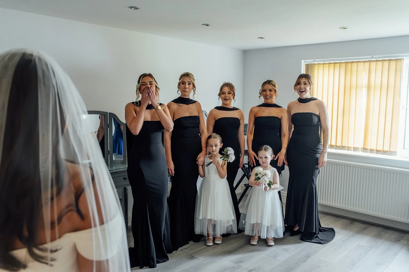 Nisha’s bridesmaids in black dresses and two young flower girls react with smiles and surprise as they see her ready in her wedding dress and veil at Batch Country House in Somerset, UK, before the ceremony, photographed by Just James Weddings.