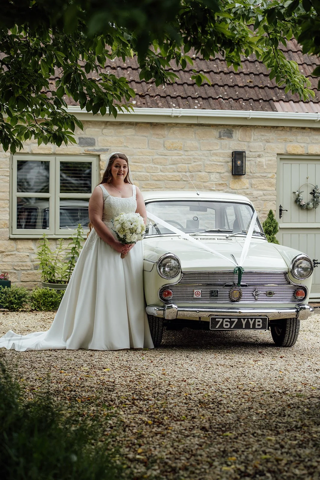 Elegant bridal portrait of Louise & Jon’s wedding at Wick Farm Bath in Somerset, UK, with the bride holding a white bouquet beside a vintage car, stone cottage backdrop, soft veil detail, and classic countryside wedding styling in the driveway.