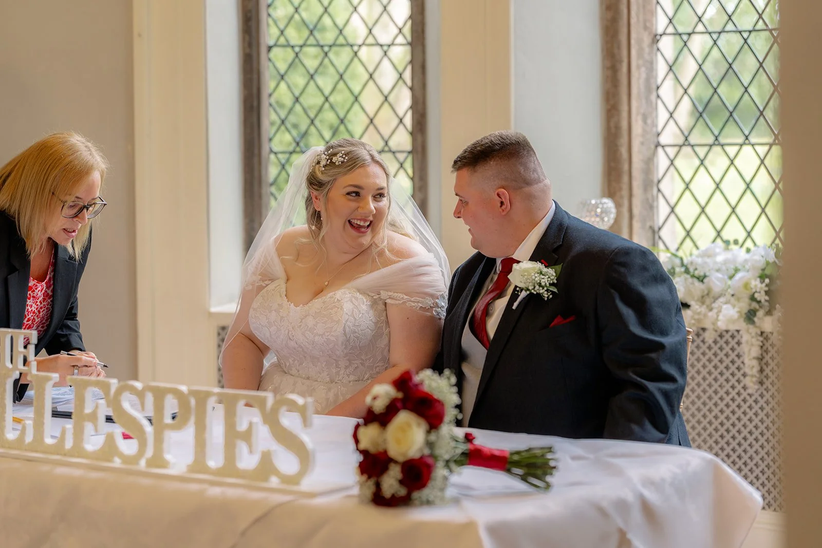 Becky & Rob sit together signing the marriage register at Clearwell Castle in Gloucestershire, UK. The bride smiles toward the groom beside the ceremony table with bouquet and flowers, captured in natural Clearwell Castle wedding photography.