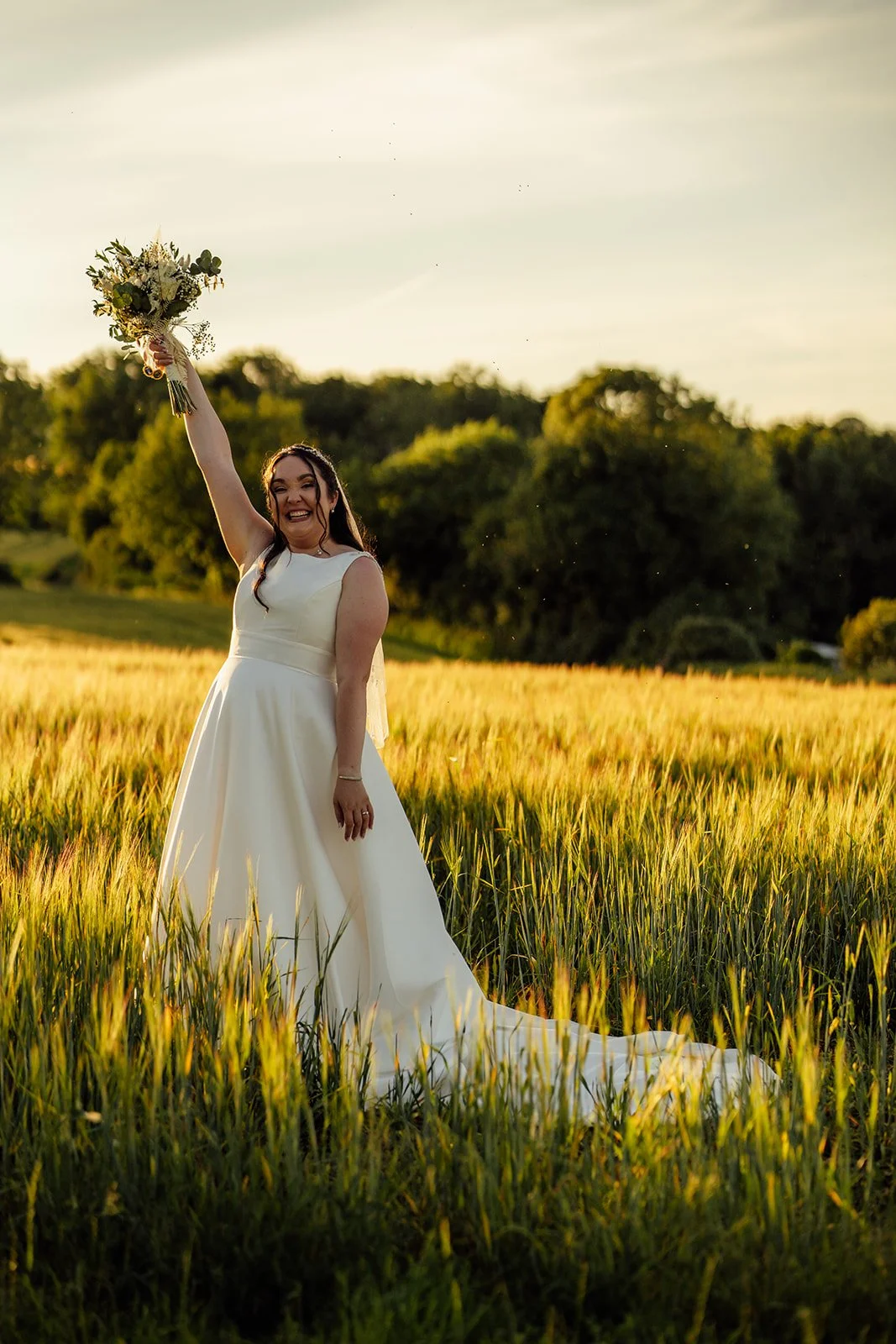 Chloe raises her wedding bouquet while standing in a sunlit field during her wedding to Armani at The Barn at Upcote in Gloucestershire, photographed by Just James Weddings.