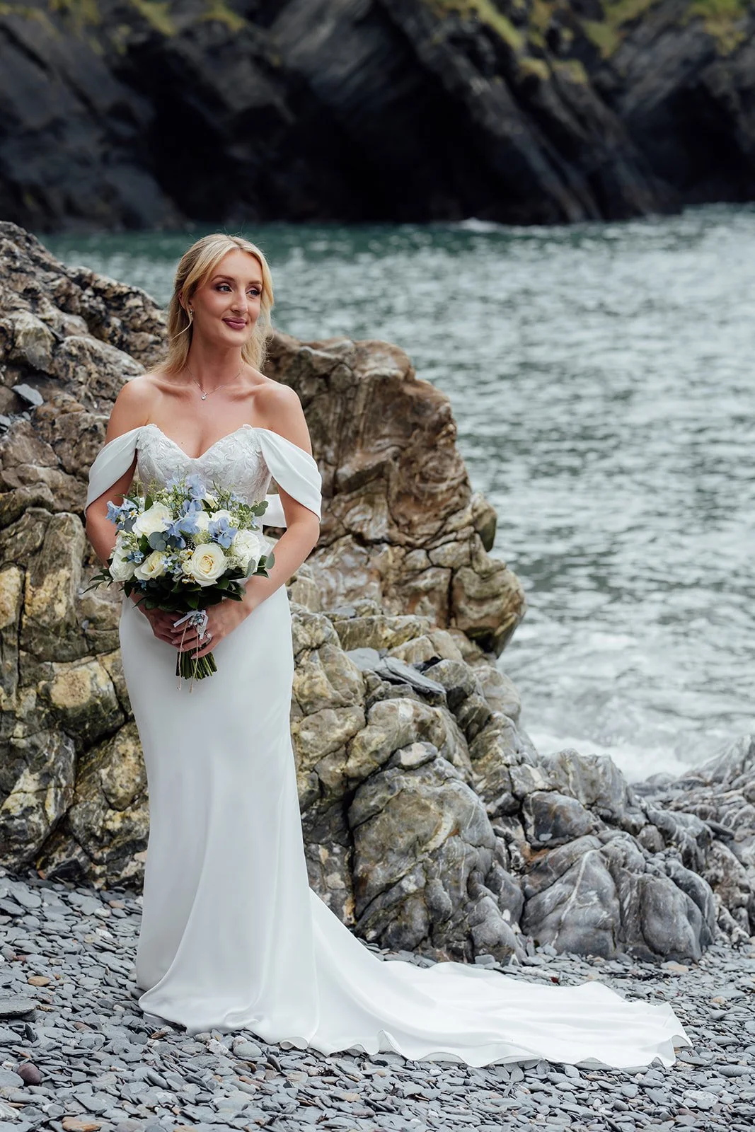 Bridal portrait of Amy holding her white and blue bouquet beside the rocks and sea near Sandy Cove Hotel in North Devon, UK.