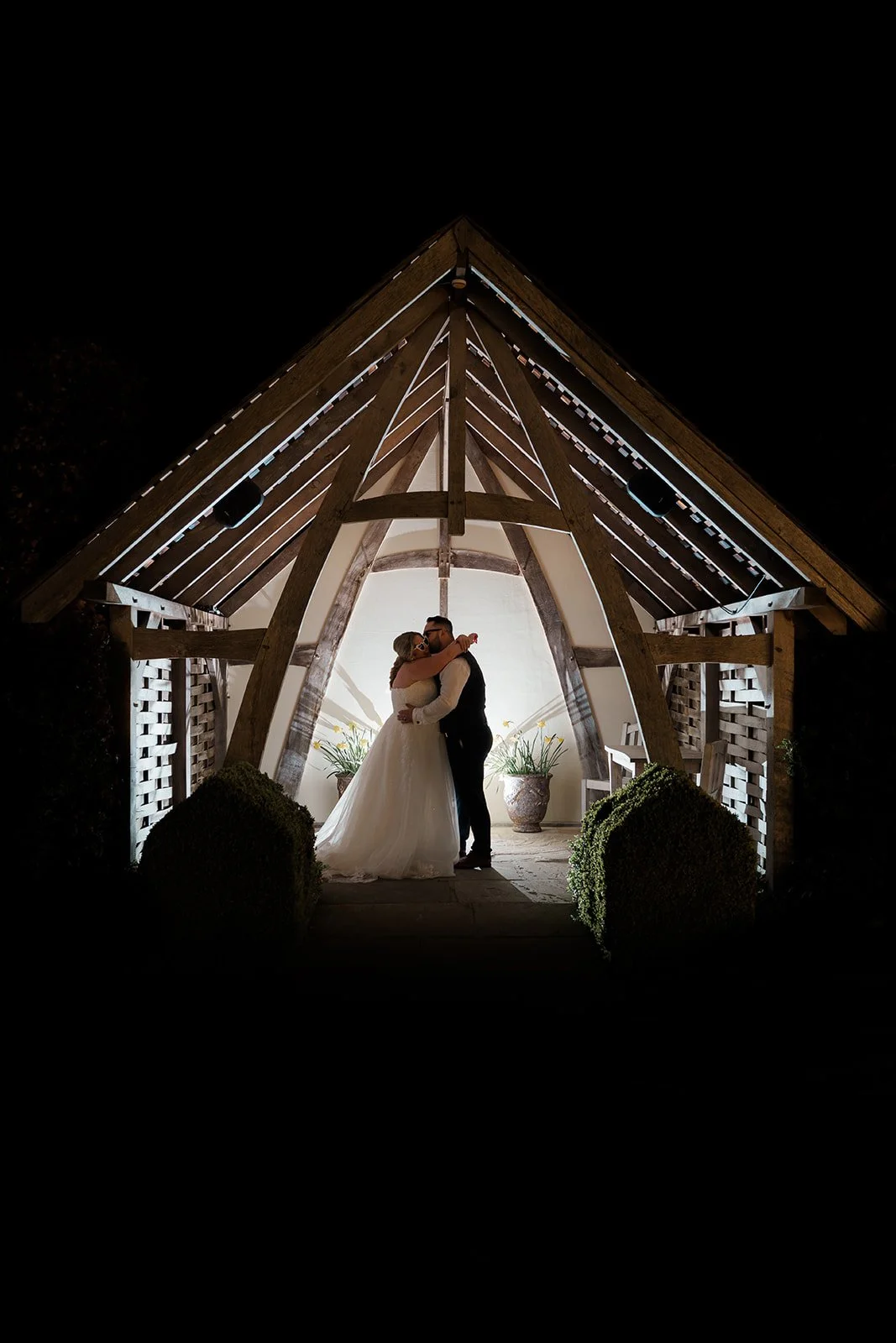 Silhouetted portrait of Siobhan & Nicholas kissing beneath the lit wooden pavilion at Kingscote Barn in the Cotswolds, UK, at night, a dramatic countryside wedding portrait with glowing light, dark sky and rustic barn venue architecture.