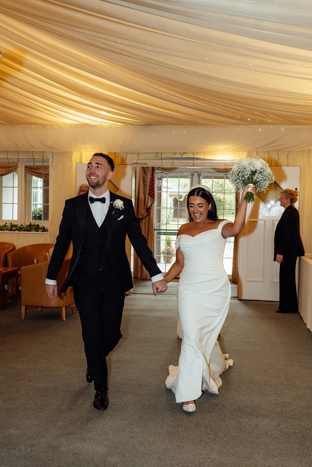 Nisha and Jack enter their wedding reception at Batch Country House in Somerset, UK, smiling and holding hands as Nisha raises her white bouquet beneath the softly draped ceiling and warm reception lighting, photographed by Just James Weddings.