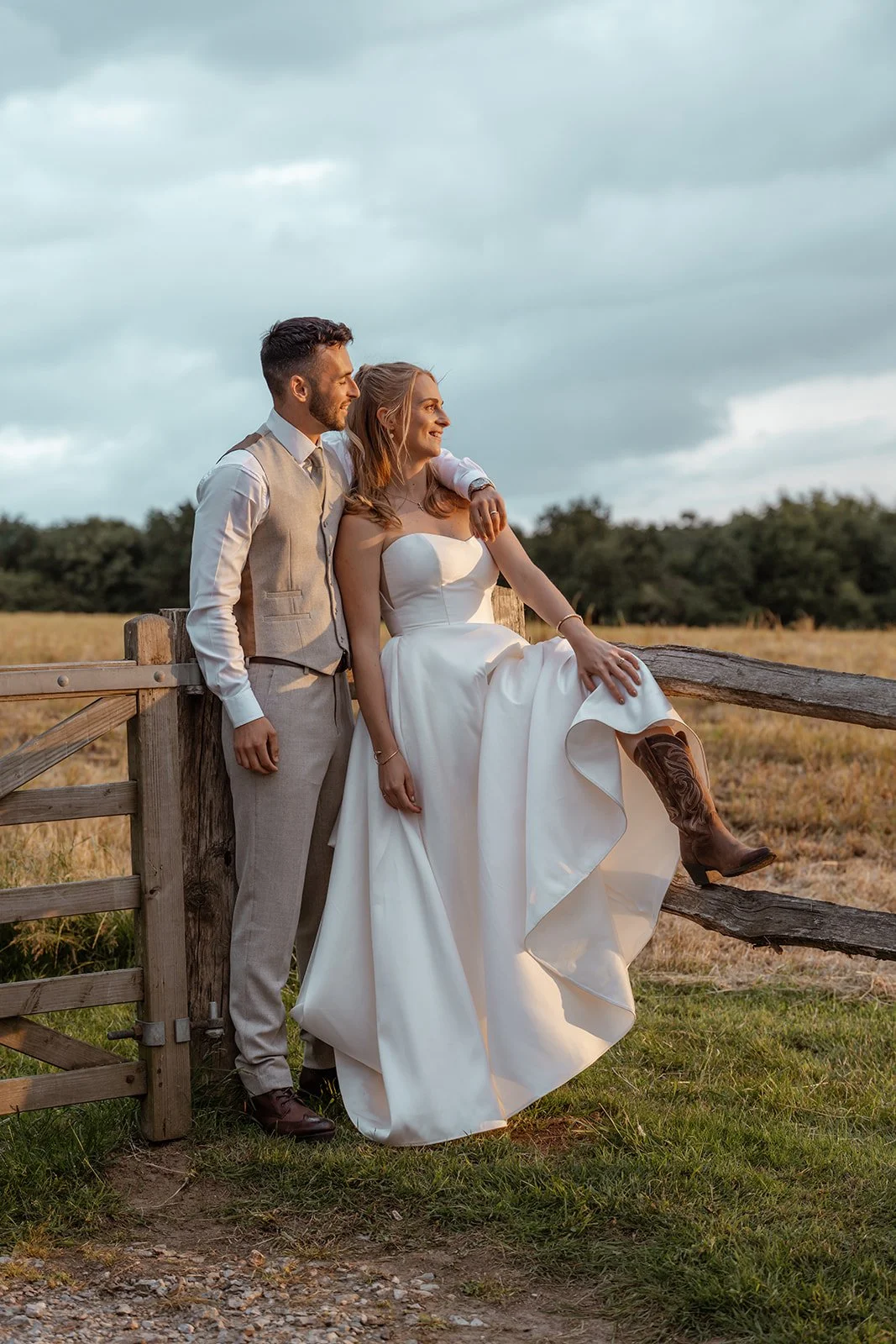 Louise & Christian sitting together on a wooden field gate at The Cider Barn in Somerset, with the bride wearing cowboy boots, photographed by Just James Weddings for relaxed countryside wedding portraits.