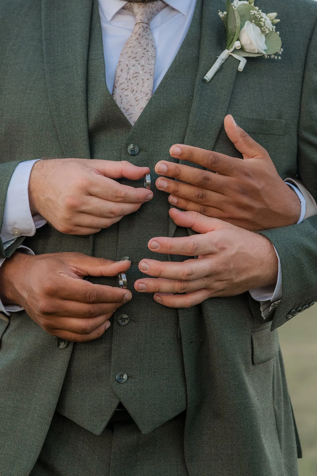 Close-up of Aaron & Jack showing their wedding rings against Jack’s green suit and white buttonhole at Rock Village Hall in Worcestershire, photographed by Just James Weddings.