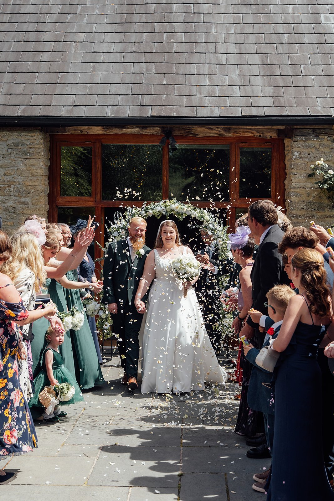 Confetti exit for Louise & Jon’s wedding at Wick Farm Bath in Somerset, UK, with the couple walking hand in hand beneath a floral arch as guests celebrate outside the stone barn, creating a bright, joyful summer wedding ceremony moment.