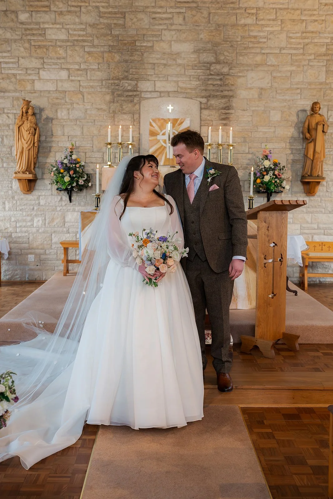Emily & Josh stand together at the altar after their ceremony at Coombe Lodge Blagdon, Somerset, UK, smiling beside pastel wedding flowers inside the church. This elegant portrait captures their just-married moment in Somerset in soft light.