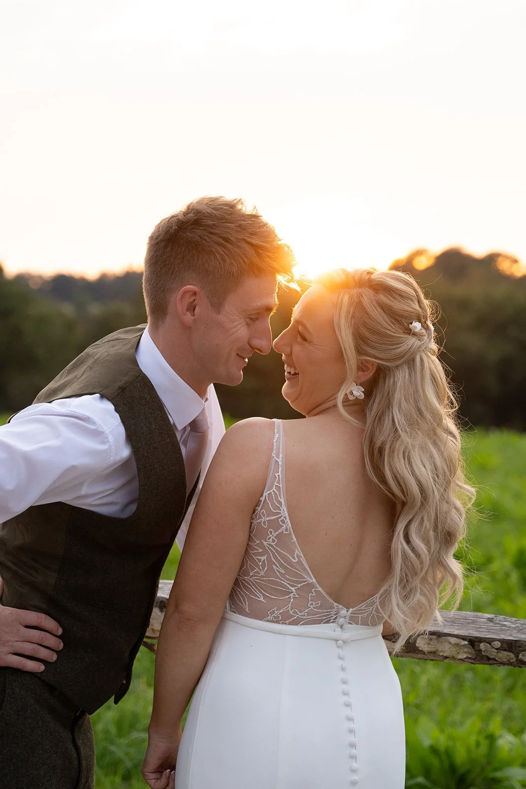 Abi & Josh laughing together at sunset at The Cider Barn in Somerset, with warm light behind them and the bride’s back detail visible, photographed by Just James Weddings for relaxed countryside wedding portraits.